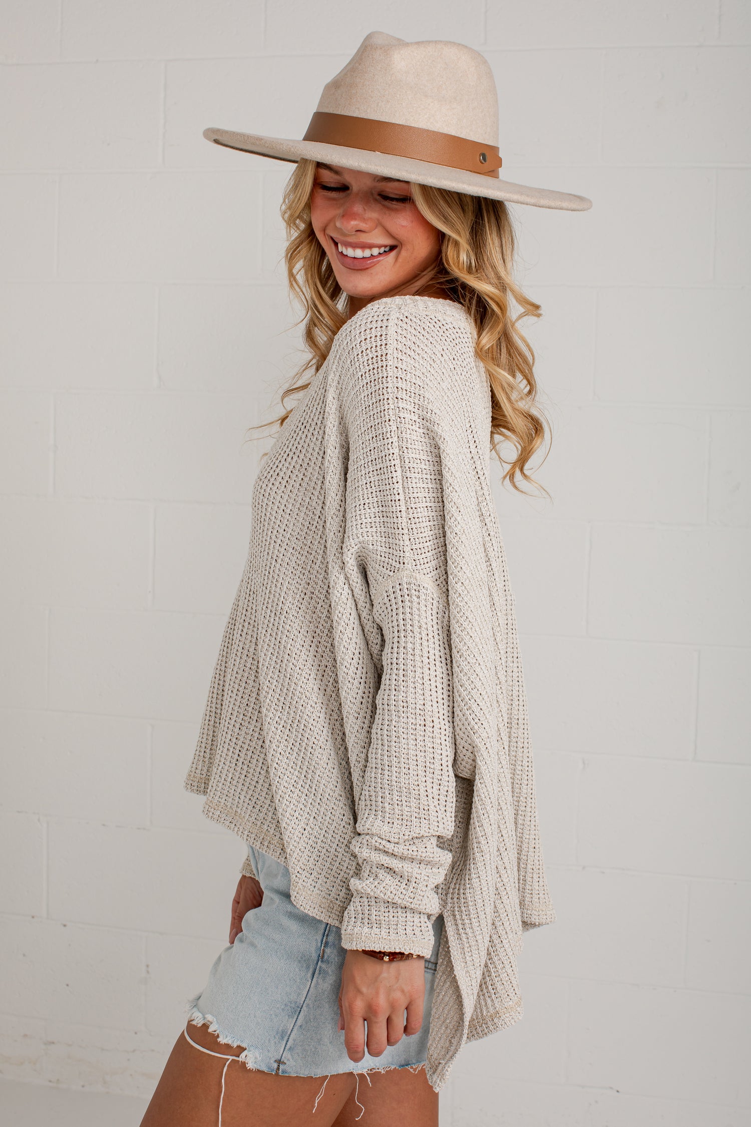 A woman wearing a cream-colored, textured knit long-sleeve poncho sweater, a wide-brimmed beige hat with a brown band, and light-wash denim shorts, posing against a white brick wall.