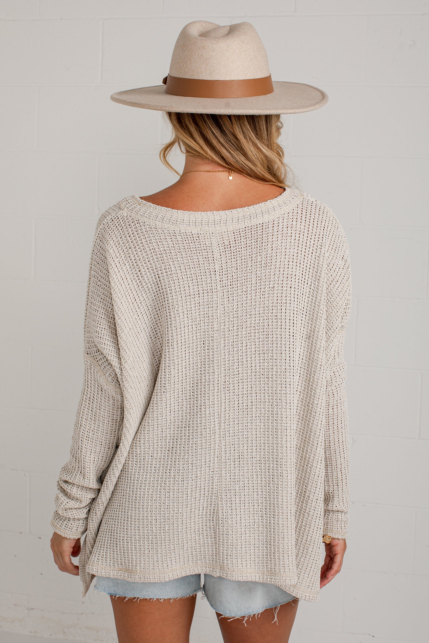A woman shown from the back wearing a cream-colored waffle-knit long-sleeve sweater, a beige wide-brim fedora hat with a brown band, and light-wash denim shorts against a white brick wall.