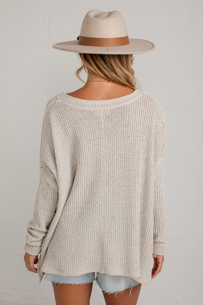A woman shown from the back wearing a cream-colored waffle-knit long-sleeve sweater, a beige wide-brim fedora hat with a brown band, and light-wash denim shorts against a white brick wall.