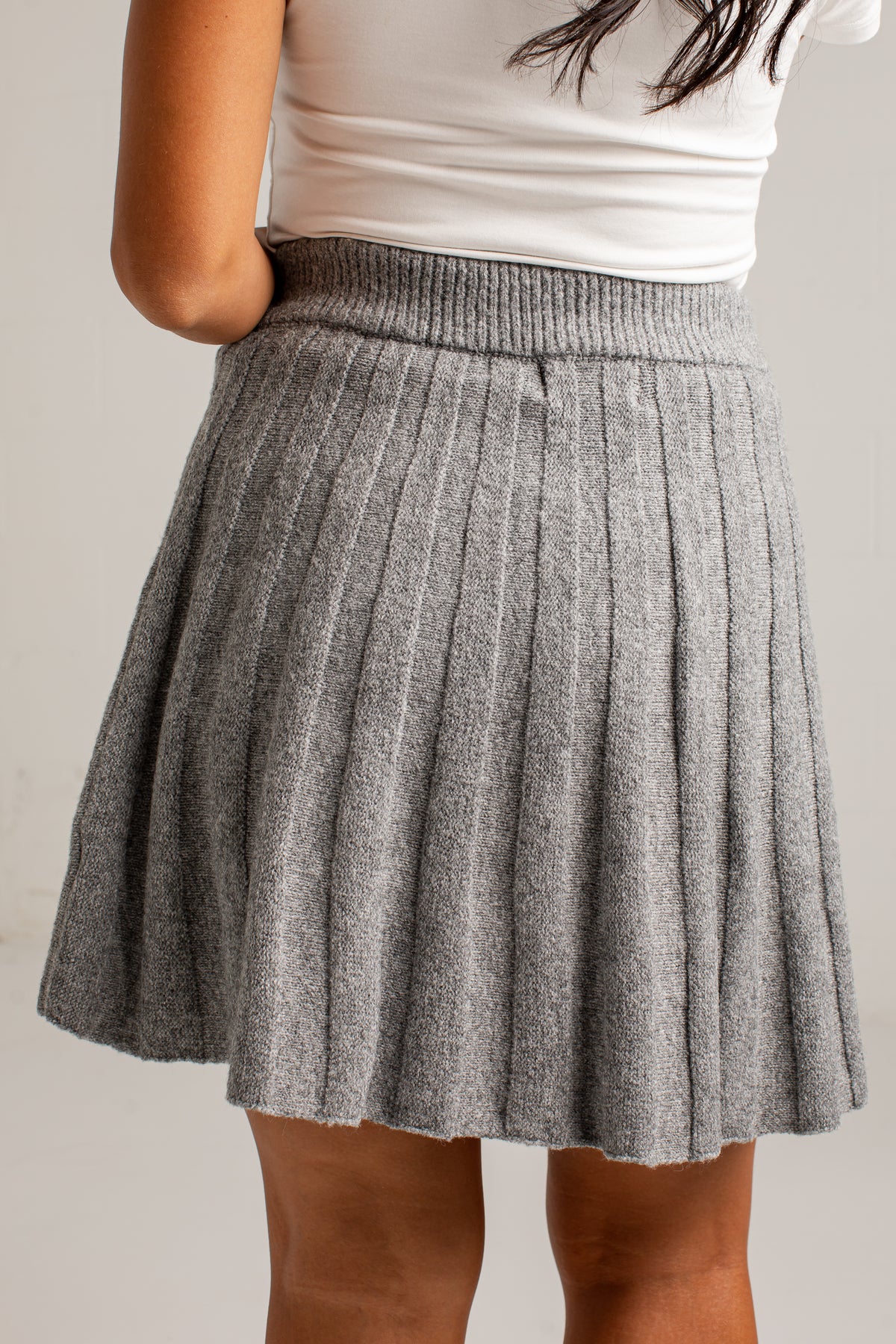 A gray pleated knit mini skirt worn by a model, paired with a white ribbed top, shown from the back against a plain light background.