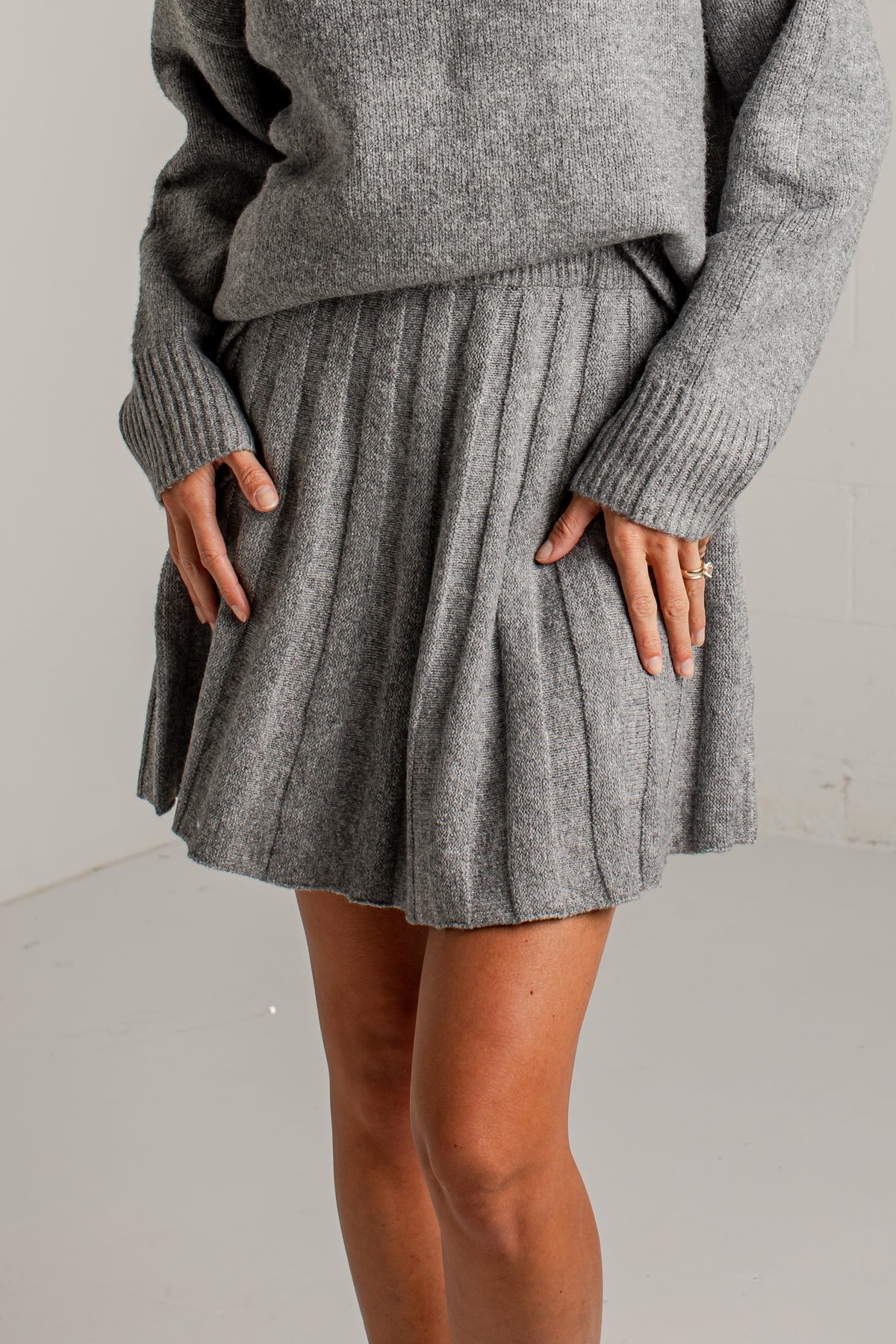 A person wearing a gray ribbed knit sweater and matching pleated mini skirt, standing against a plain light gray wall.