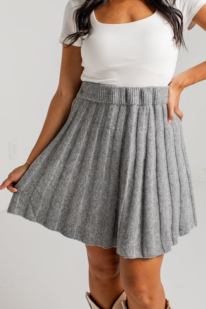 A woman wearing a gray pleated knit mini skirt with a ribbed waistband, styled with a white scoop-neck top and beige boots against a plain light gray background.