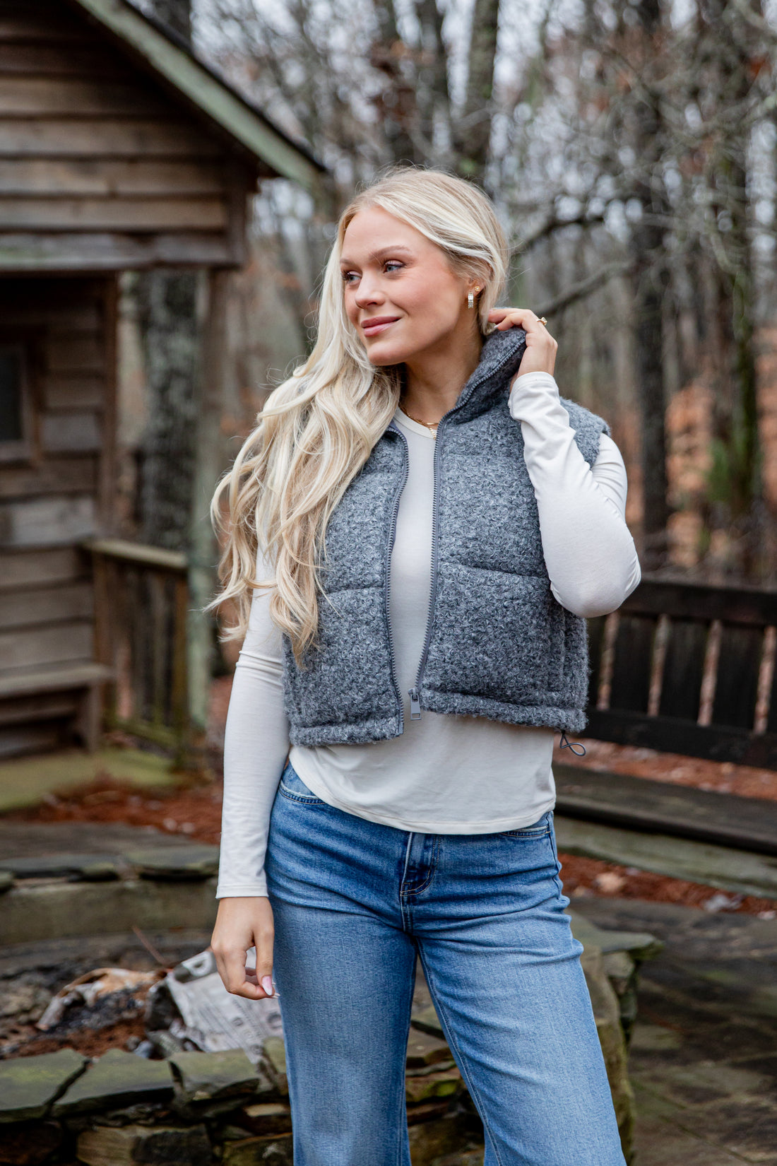 Woman wearing a blue puffer vest and jeans standing outdoors near a wooden cabin.
