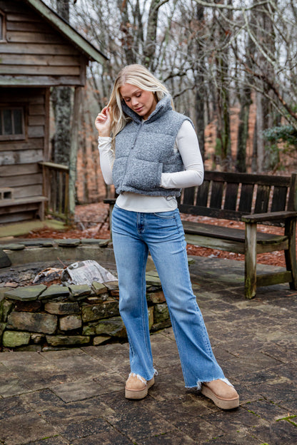 Woman wearing a gray puffer vest and blue jeans standing outdoors near a wooden cabin.