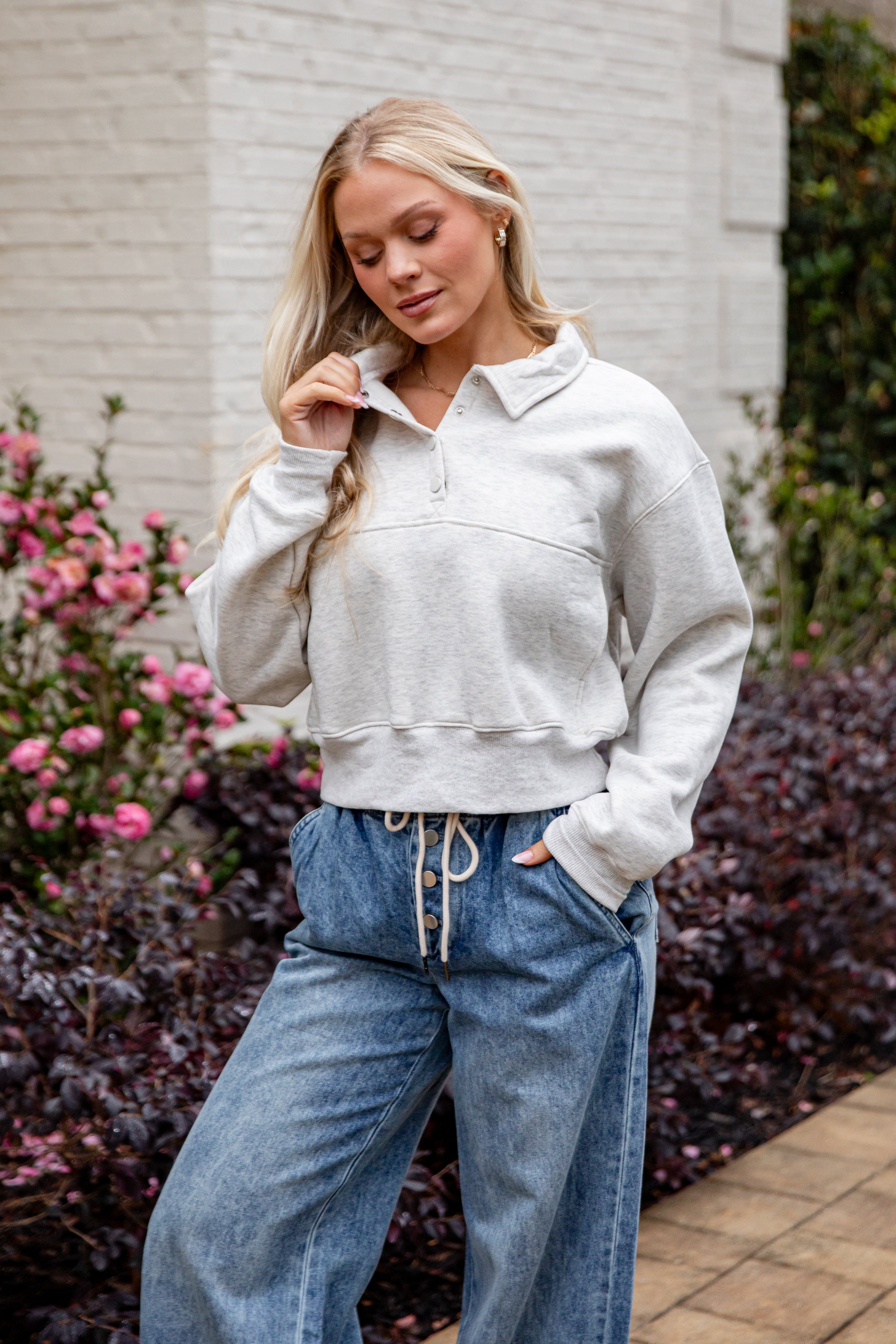 Woman wearing a light gray sweater and blue jeans standing outdoors with flowers and a building in the background.