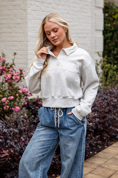 Woman wearing a light gray sweater and blue jeans standing outdoors with flowers and a building in the background.