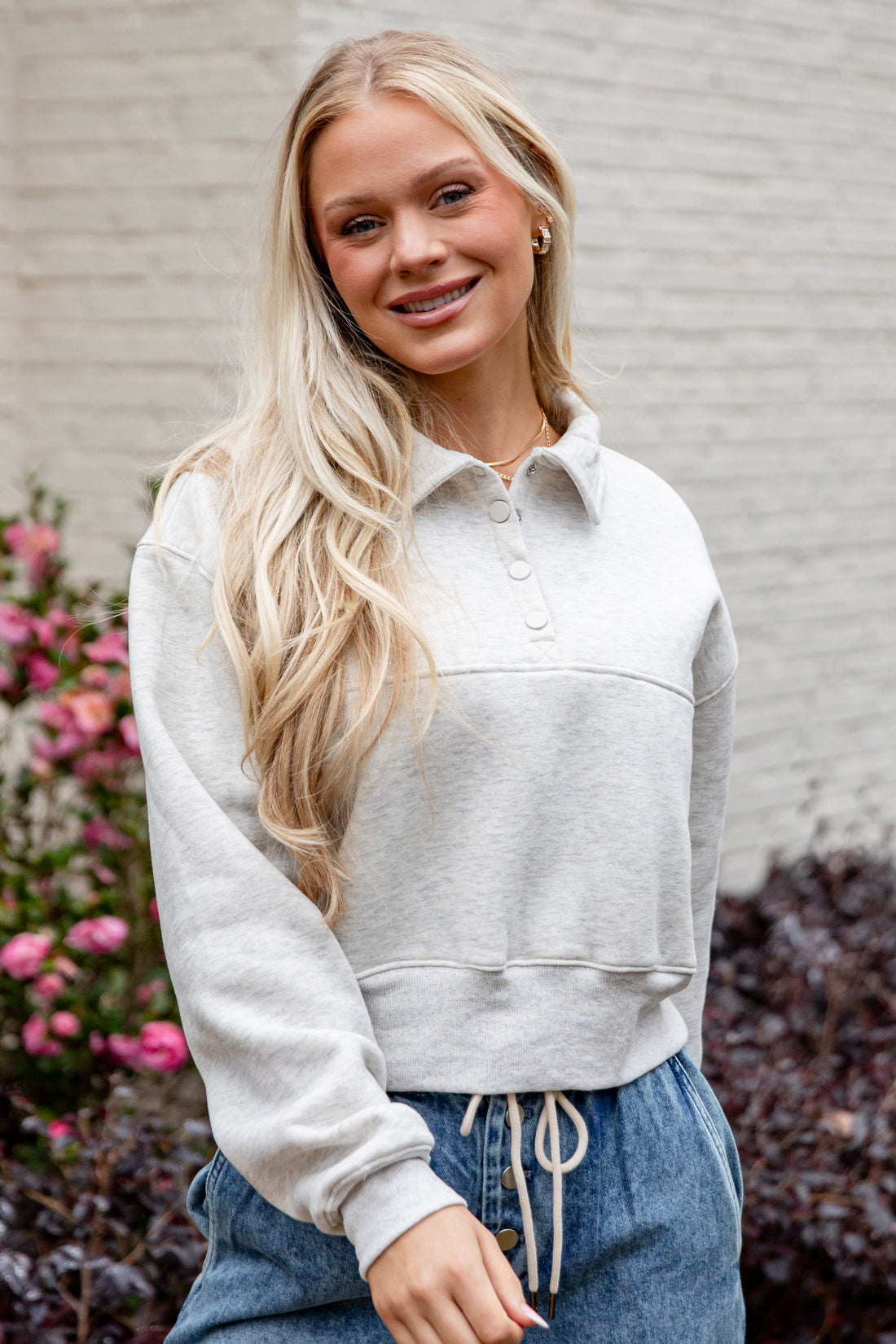 Woman wearing a light gray cropped hoodie and blue jeans standing in front of a white wall with flowers.