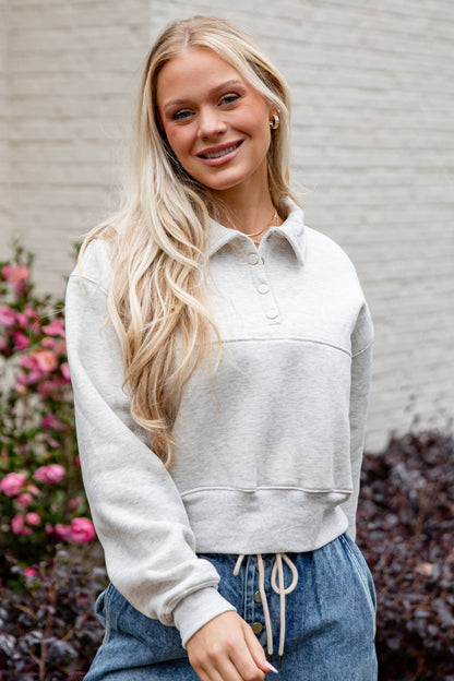 Woman wearing a light gray cropped hoodie and blue jeans standing in front of a white wall with flowers.
