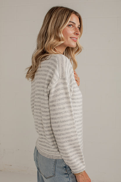 A woman with blonde hair is shown from a side-back angle wearing a light gray and white horizontally striped knit sweater and light blue jeans against a white brick wall.