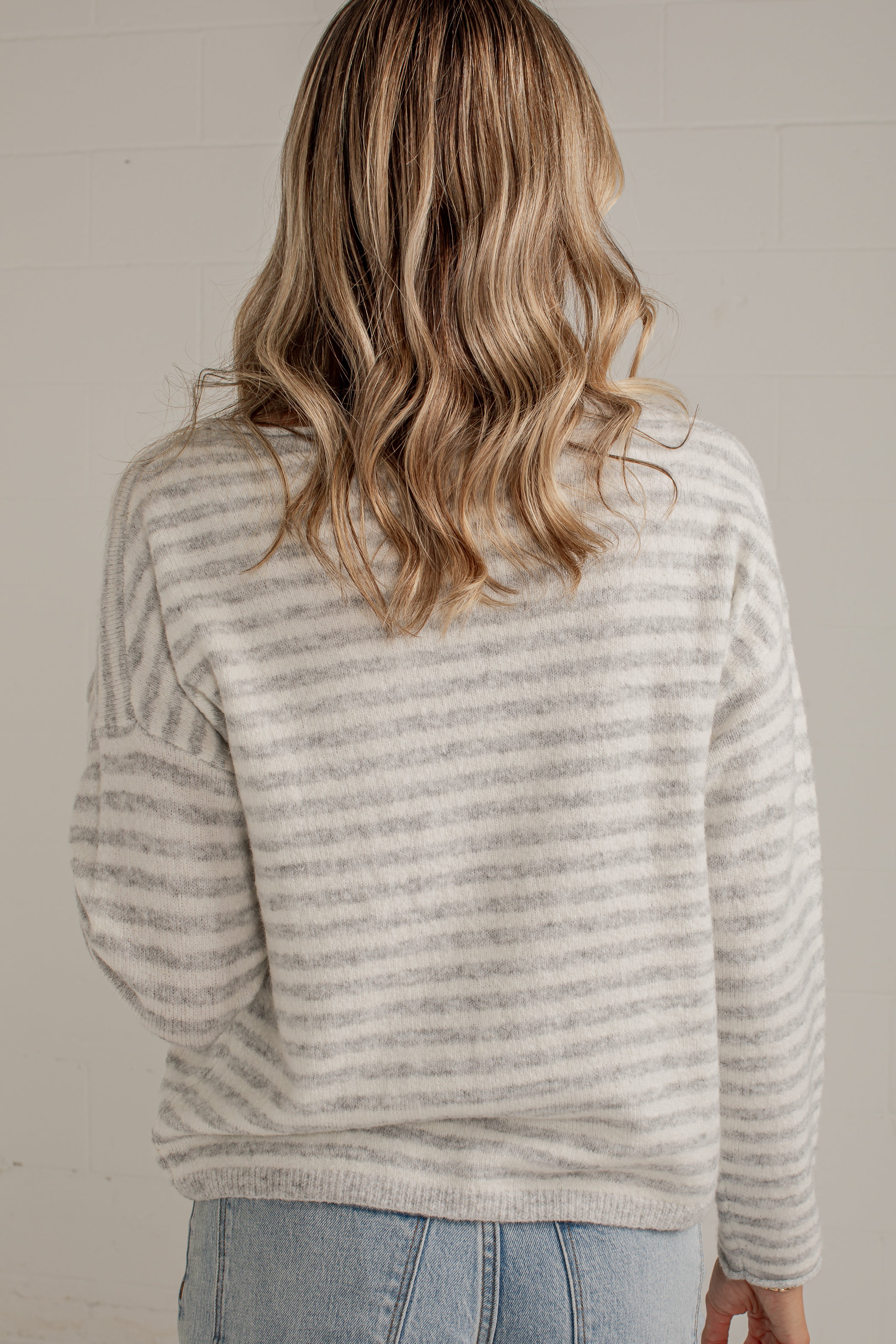 A woman viewed from behind wearing a light gray and white horizontally striped knit sweater and light blue jeans against a neutral off-white wall.