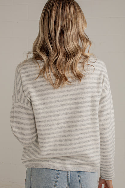 A woman viewed from behind wearing a light gray and white horizontally striped knit sweater and light blue jeans against a neutral off-white wall.