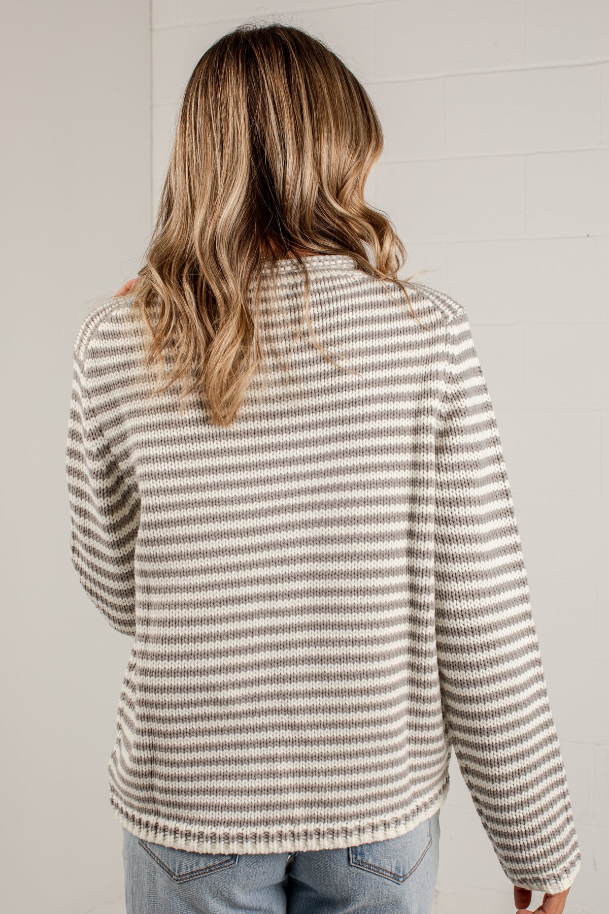 A woman viewed from behind wearing a long-sleeve knit sweater with thin horizontal beige and gray stripes, paired with light blue jeans, against a light gray brick wall.