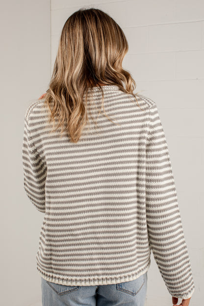 A woman viewed from behind wearing a long-sleeve knit sweater with thin horizontal beige and gray stripes, paired with light blue jeans, against a light gray brick wall.