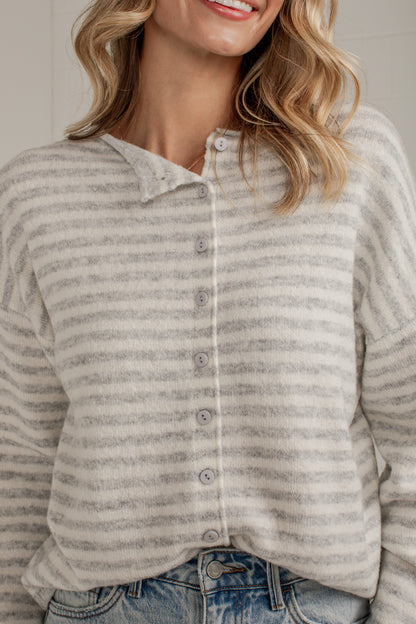 A close-up of a woman wearing a light gray and white heathered striped long-sleeve button-up henley shirt with a collar, paired with blue jeans.