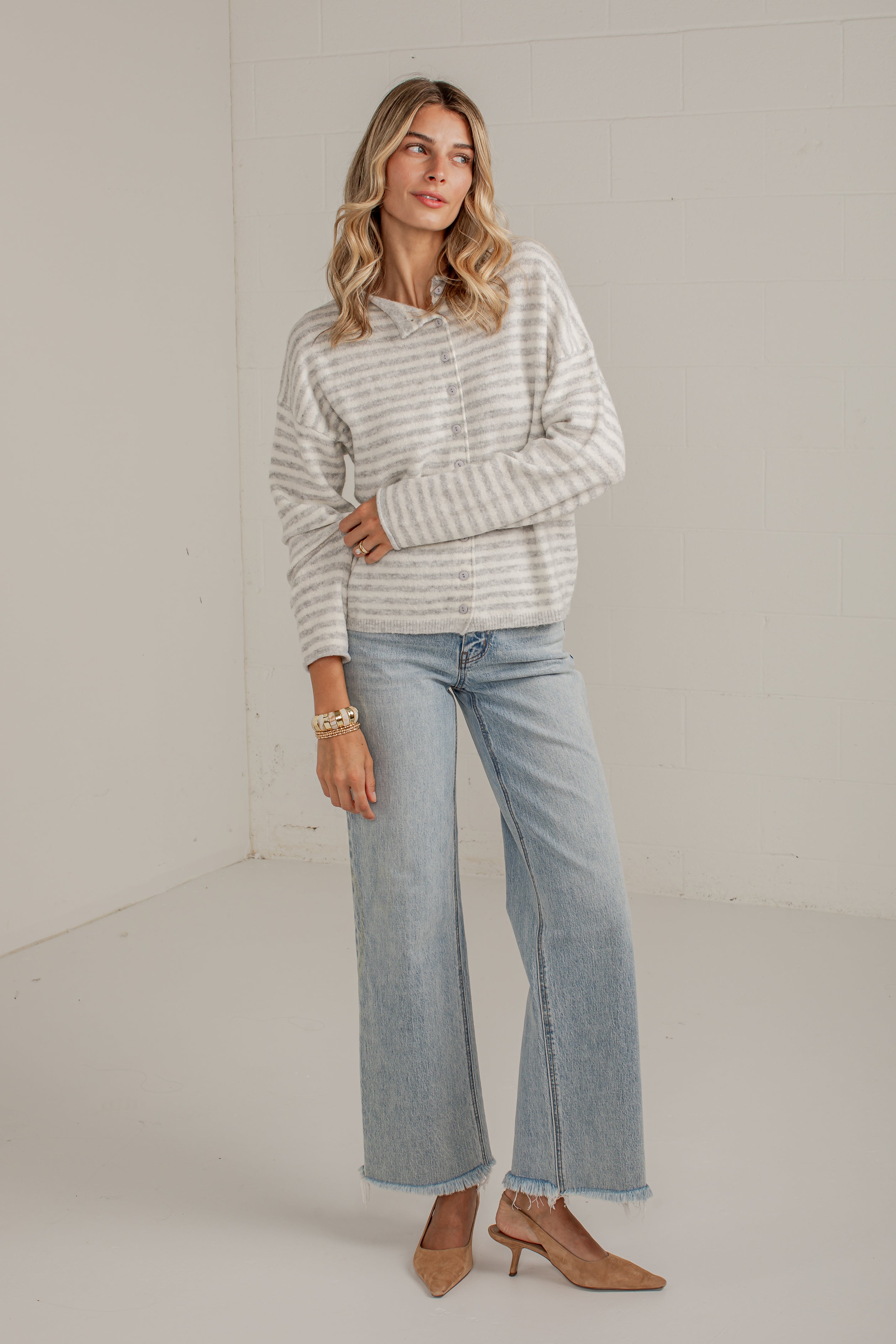 A woman modeling a light gray and white striped zip-up hoodie with wide-leg light-wash blue jeans and tan heeled sandals, standing against a plain off-white studio wall.