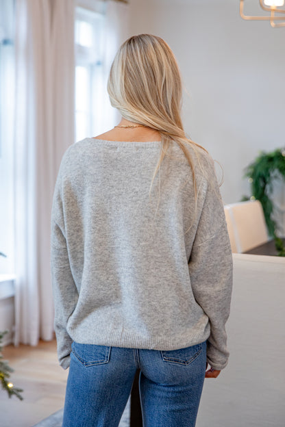 Person wearing a gray sweater and blue jeans in a room with white walls and a plant.