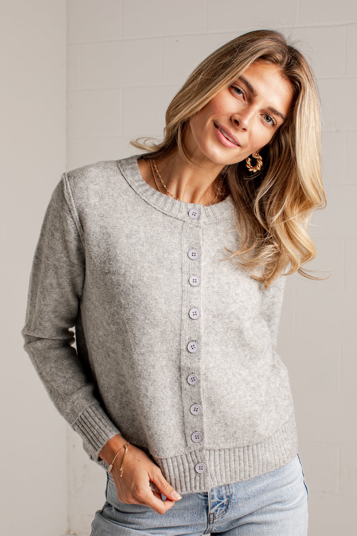 A woman wearing a light gray heathered knit cardigan sweater with a button-up front, paired with light-wash jeans, standing against a light gray brick wall.