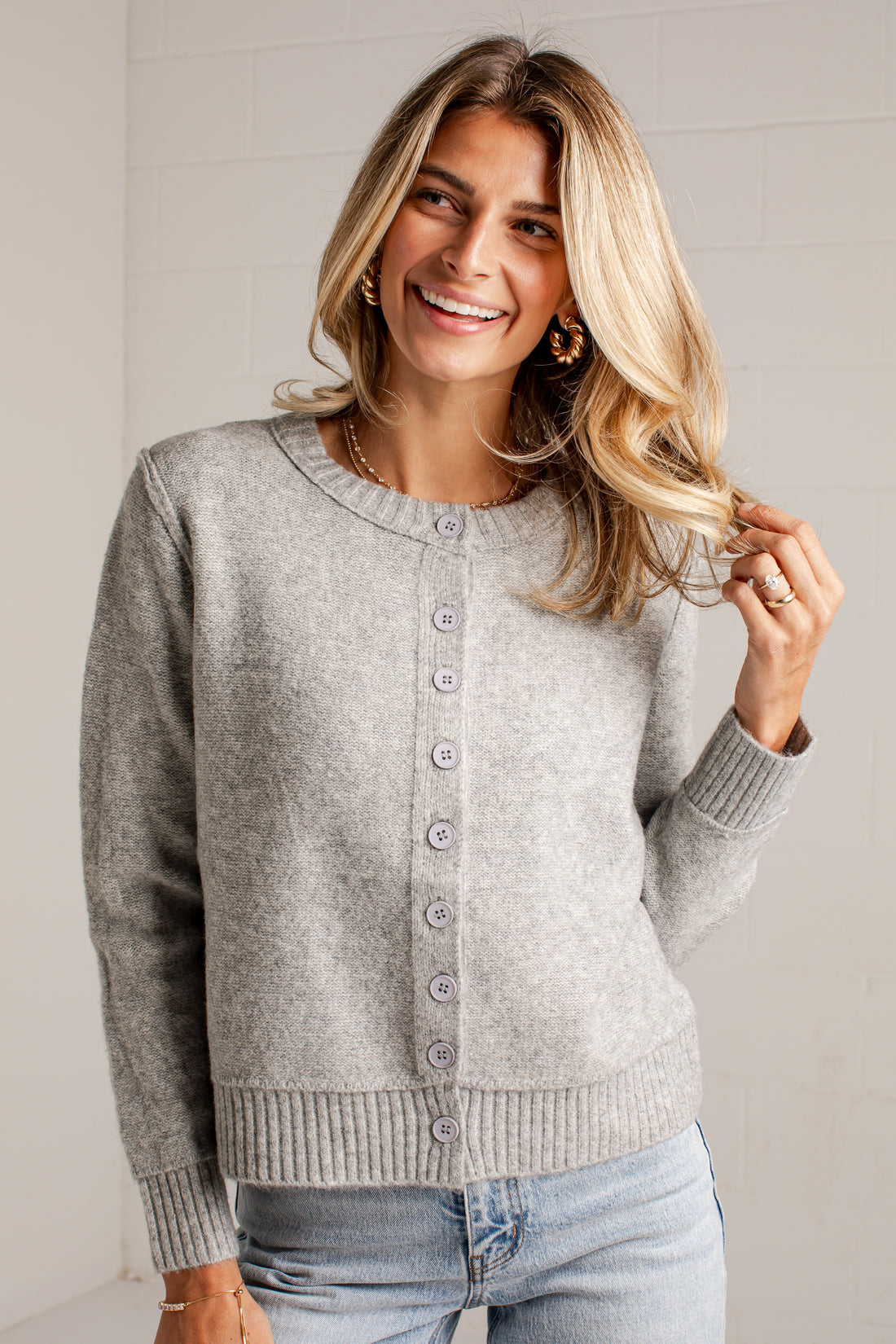 A woman wearing a light gray knit cardigan sweater with a button-up front, paired with light blue jeans, standing against a white brick wall.