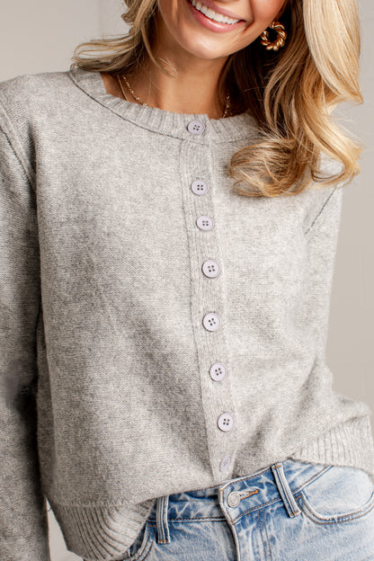 A close-up of a woman wearing a light gray heather knit cardigan with a button-front closure and light gray buttons, paired with blue jeans.