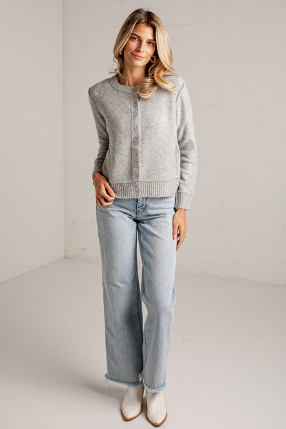 A woman modeling a light gray knit cardigan sweater with front buttons, paired with light-wash wide-leg jeans and white ankle boots, standing in a bright white studio corner.