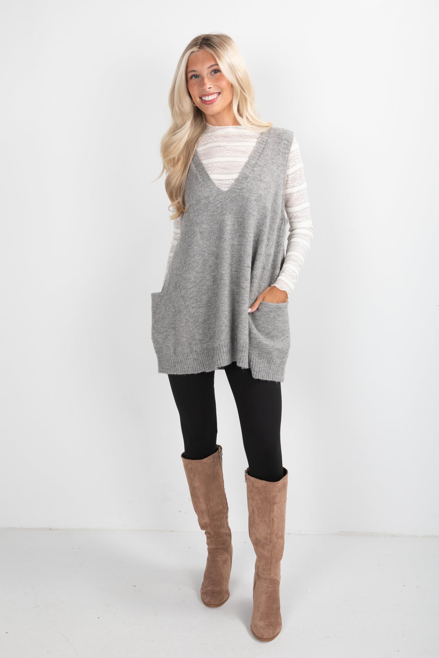 Woman wearing a gray sleeveless sweater, black leggings, and brown knee-high boots on a white background