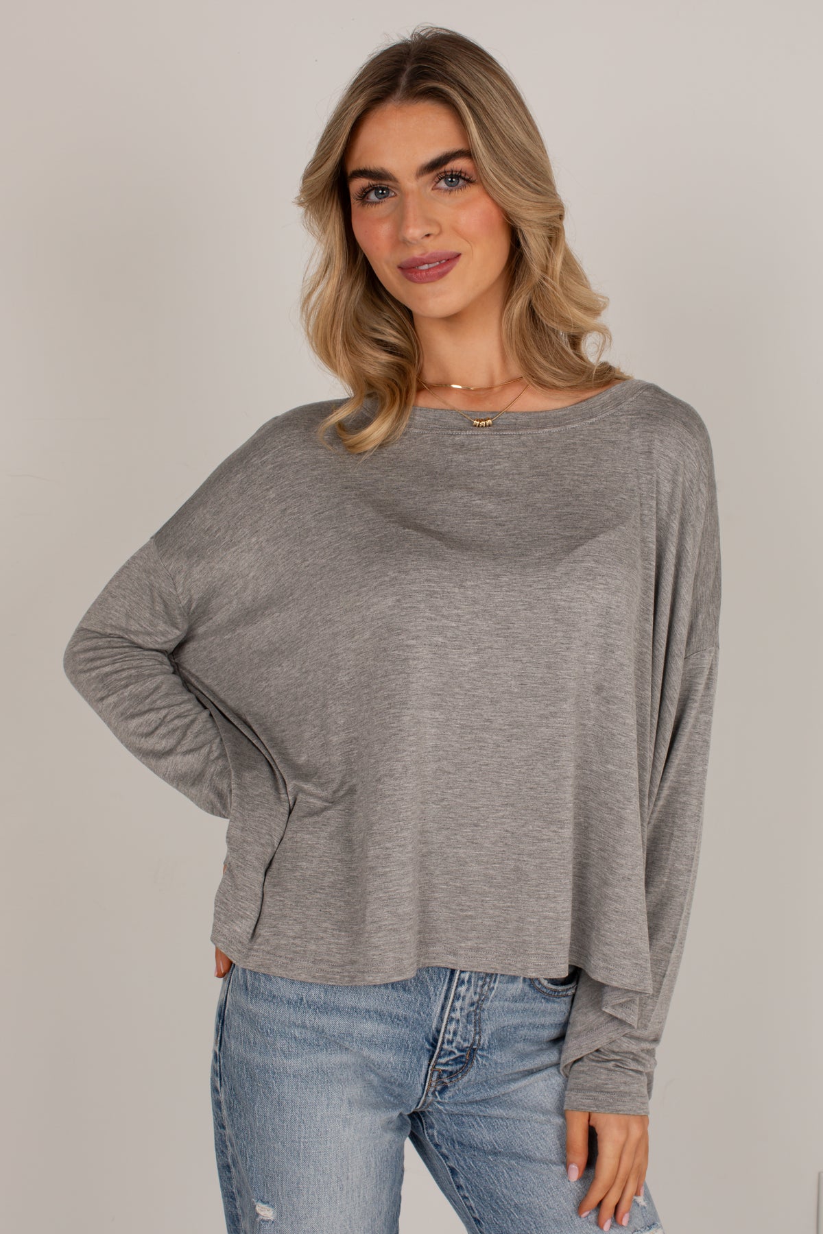 A woman wearing a heather gray long-sleeve top with a wide boat neckline and relaxed fit, paired with light blue jeans, against a plain light gray studio background.