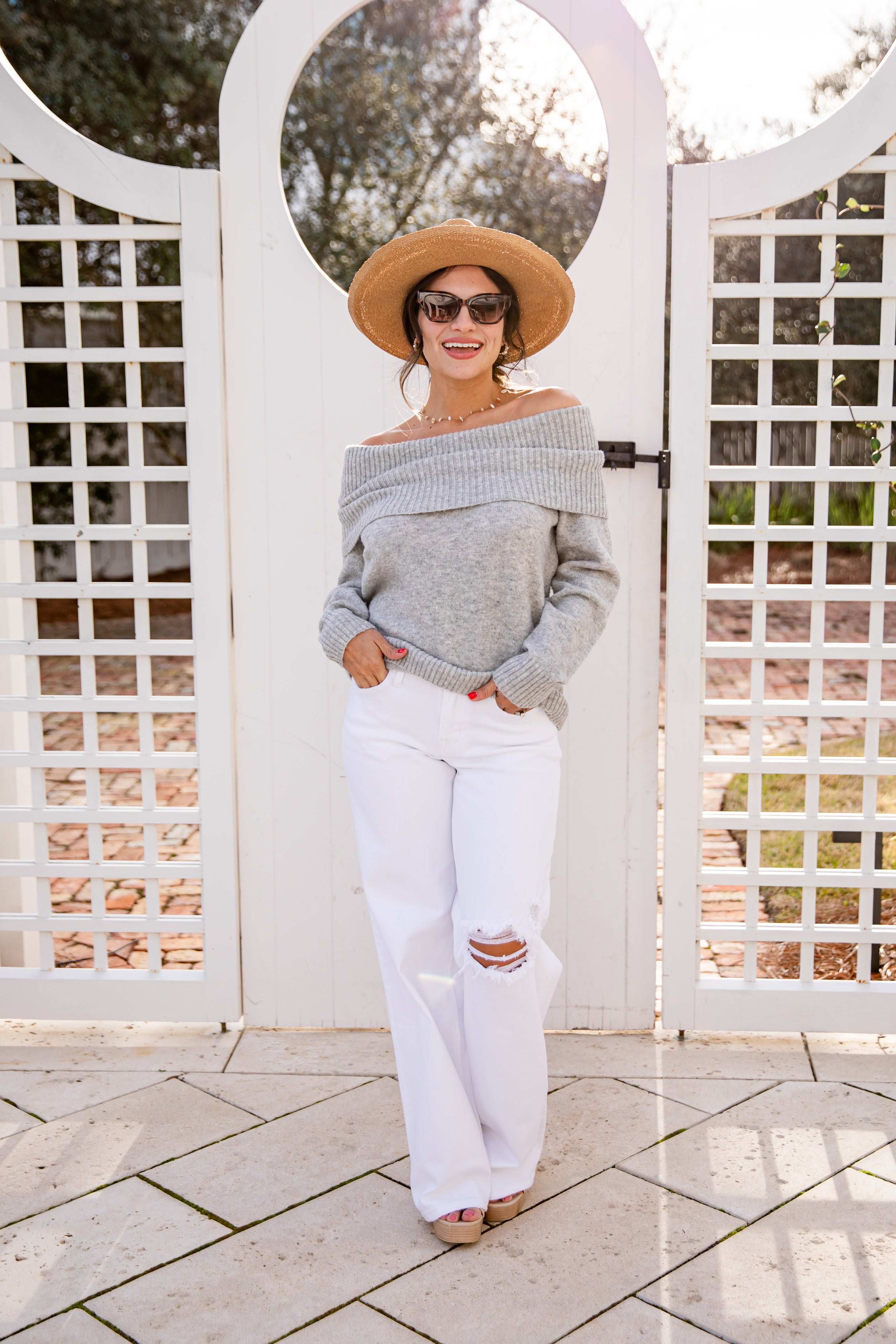 Woman wearing a gray off-shoulder sweater, white pants, and a straw hat standing in front of a white lattice gate.