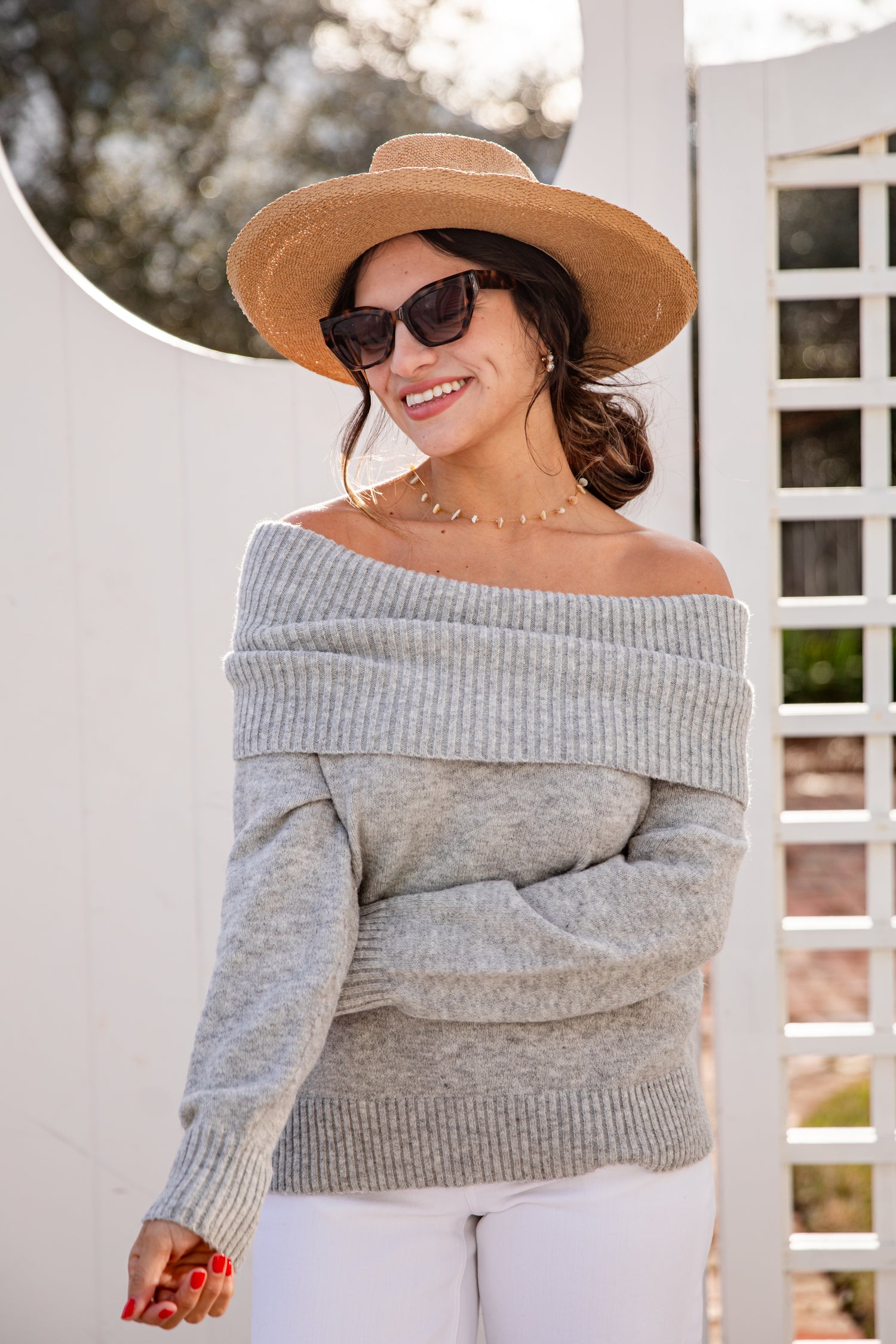 Woman wearing a gray off-shoulder sweater, white pants, and a wide-brimmed hat outdoors.
