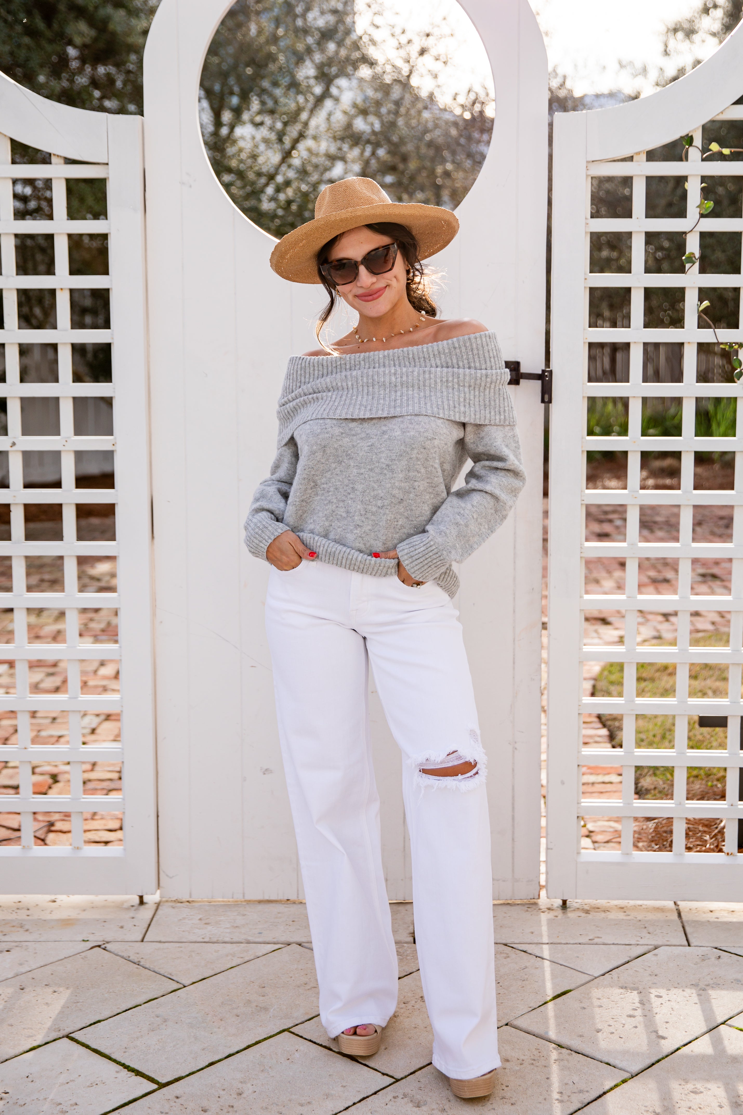 Woman wearing a gray sweater and white pants standing in front of a white lattice gate.