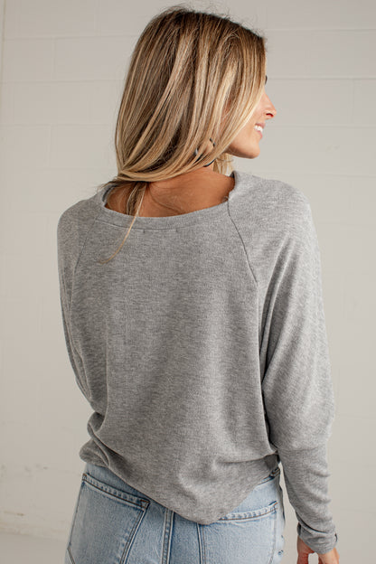 A woman shown from the back wearing a heather gray long-sleeve raglan top and light blue jeans, standing against a light off-white brick wall.