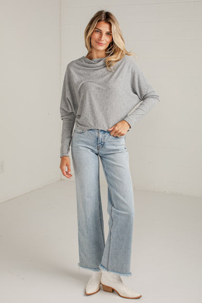 A woman modeling a light gray long-sleeve knit top and light-wash wide-leg jeans with white ankle boots, standing against a white studio wall.