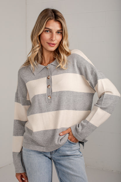 A woman wearing a gray and cream striped long-sleeve knit polo sweater with a buttoned henley collar, paired with light-wash blue jeans, standing against a light gray brick wall.