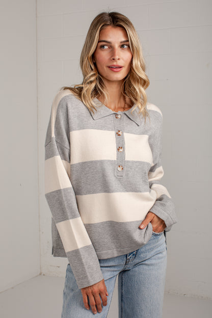 A woman wearing a gray and cream striped long-sleeve polo knit sweater with a three-button placket, paired with light-wash blue jeans, standing against a white paneled wall.