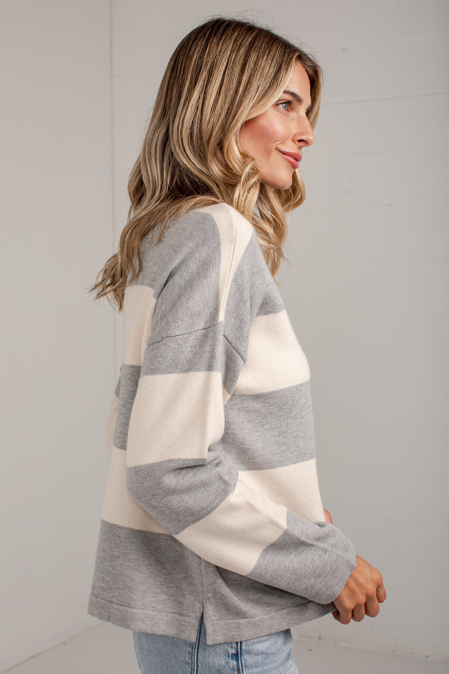A woman wearing a gray and cream color-blocked knit sweater, shown from a side profile against a light gray wall.