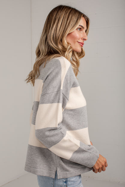 A woman wearing a gray and cream color-blocked knit sweater, shown from a side profile against a light gray wall.