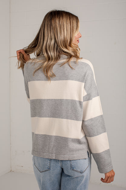 A woman shown from the back wearing a gray and cream striped long-sleeve sweater with light blue jeans, standing against a white paneled wall.