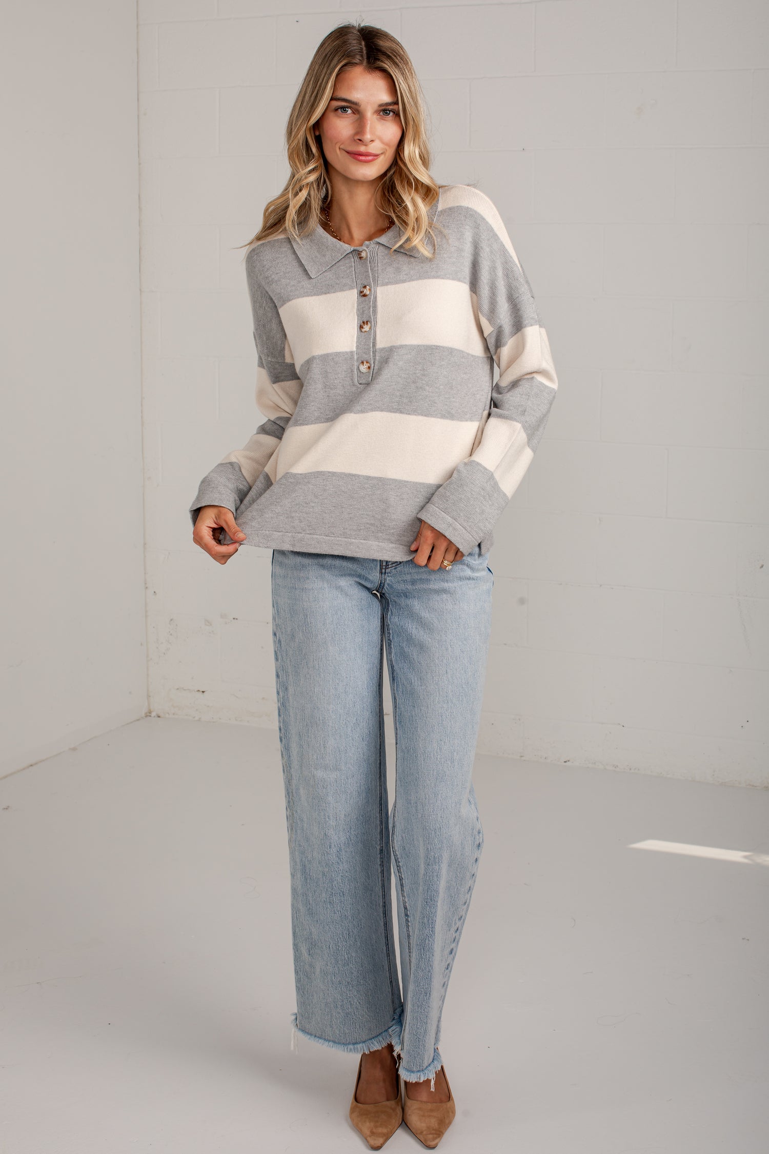 A woman modeling a gray and cream striped long-sleeve henley sweater with light-wash wide-leg jeans and brown pointed-toe heels, standing in a bright white studio corner.