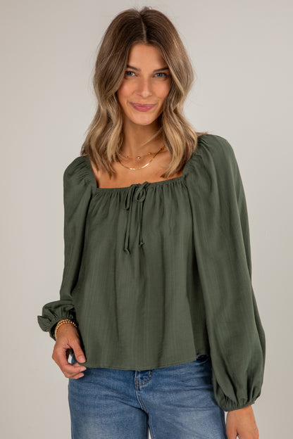 Woman wearing an olive green blouse with long sleeves and blue jeans on a plain background