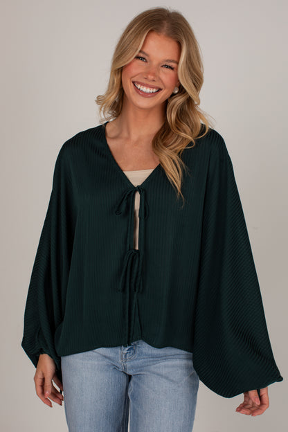 A smiling blonde woman wearing a dark forest green long-sleeve top with a tie-front keyhole detail and light-wash blue jeans, photographed against a plain light gray studio background.