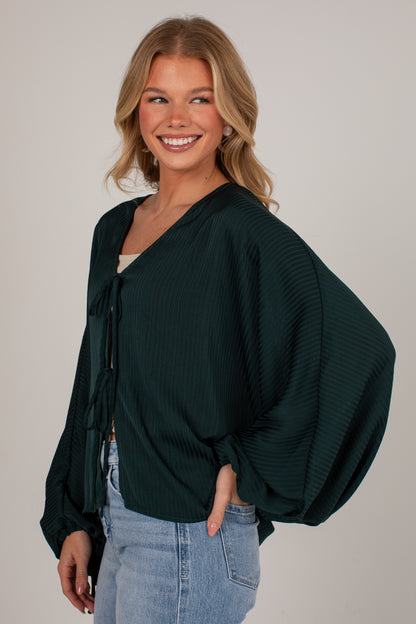 A smiling blonde woman wearing a dark forest green pleated long-sleeve blouse with balloon sleeves and a tie neckline, paired with light blue jeans, against a solid light gray background.