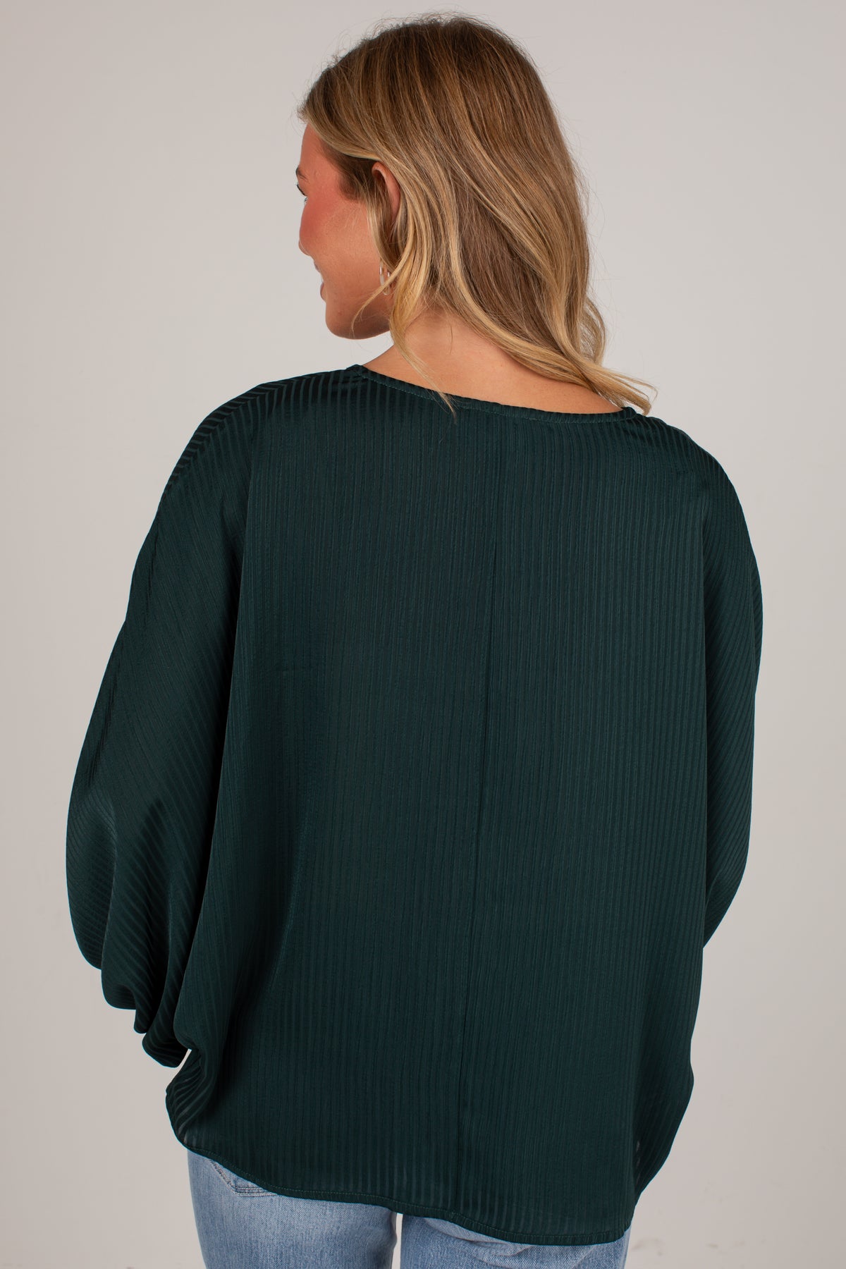 A woman viewed from behind wearing a dark forest green ribbed knit top with balloon sleeves, paired with light blue jeans, against a plain light gray background.