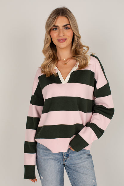 A woman modeling a pink and dark green horizontally striped long-sleeve henley sweater with a white inner placket, paired with light blue jeans, against a solid light gray background.