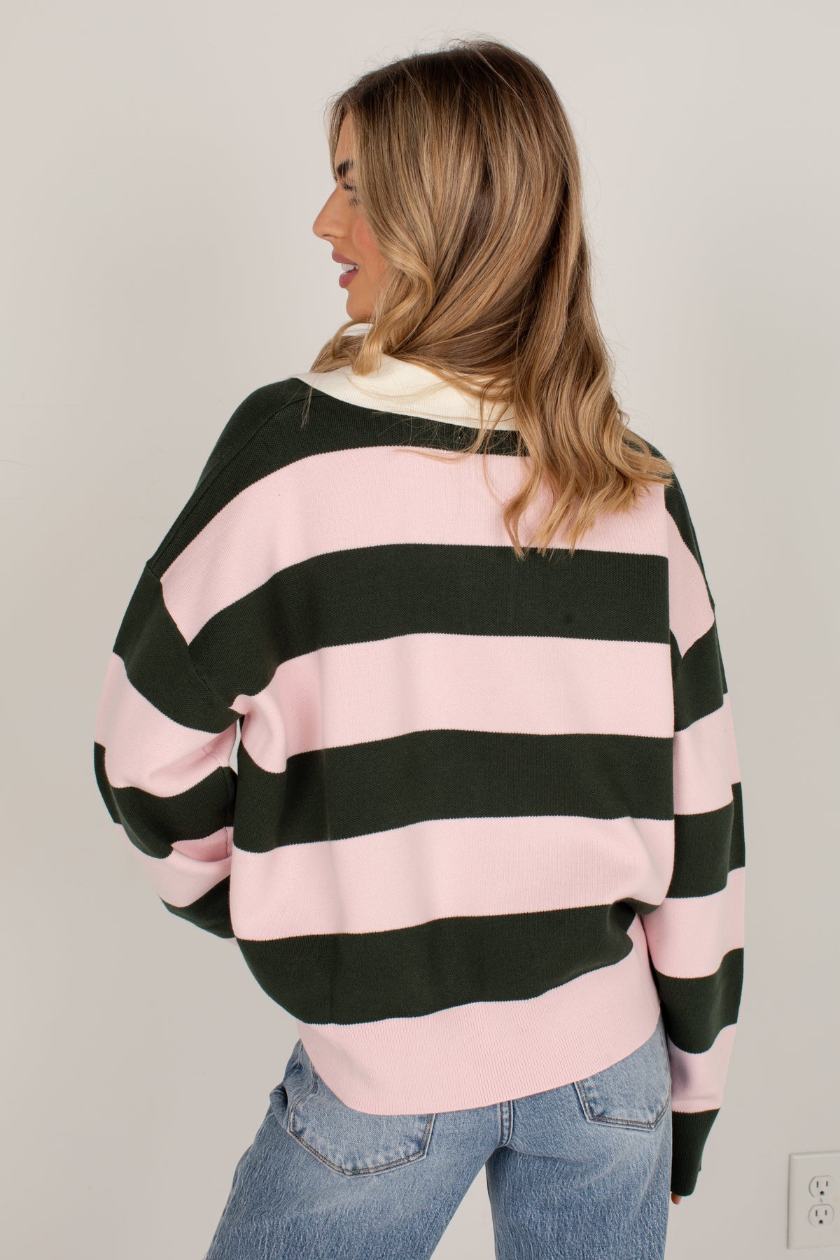 A woman shown from the back wearing a pink and dark green horizontally striped sweatshirt with a cream ribbed collar, paired with blue jeans, against a plain light gray wall.