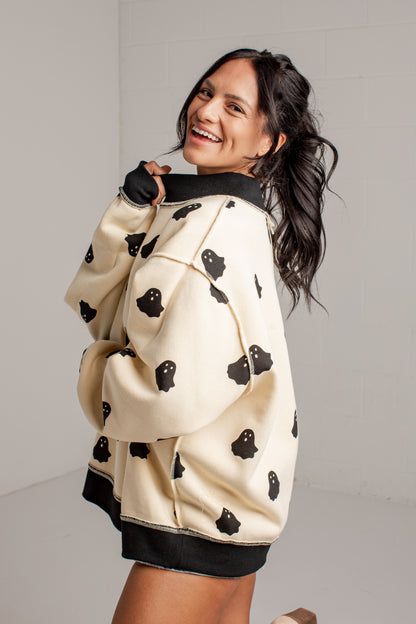 A woman wearing a cream-colored fleece hoodie with black ghost patterns and black ribbed trim, posing in a bright studio setting.