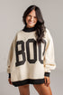 A woman wearing an oversized cream-colored knit sweater with black ribbed trim and large black block letters spelling &