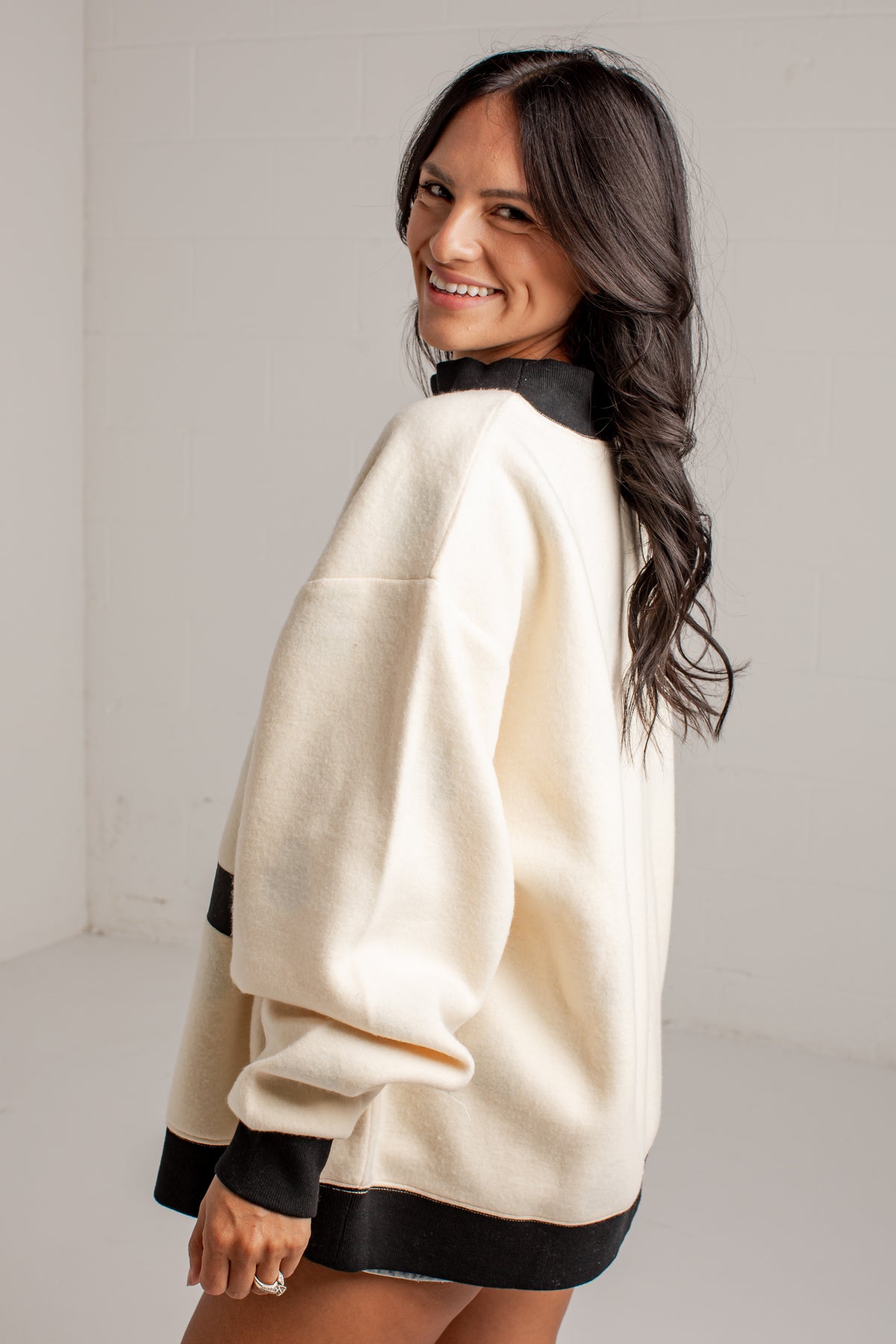 A woman wearing a cream-colored oversized sweatshirt with black ribbed trim on the collar, cuffs, and hem, photographed in a bright studio setting.