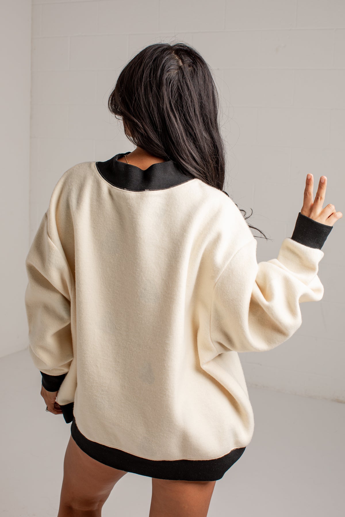 A woman shown from behind wearing an oversized cream sweatshirt with black ribbed trim at the collar, cuffs, and hem, against a plain light gray wall.