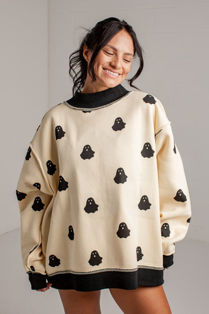 A woman wearing an oversized cream sweatshirt with black ghost patterns and black ribbed trim, smiling in a bright indoor setting.