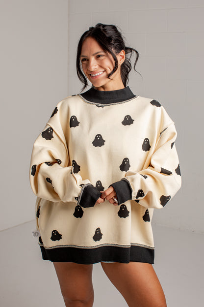 A woman wearing an oversized cream sweatshirt with black ghost patterns and black ribbed trim, standing against a light gray wall.