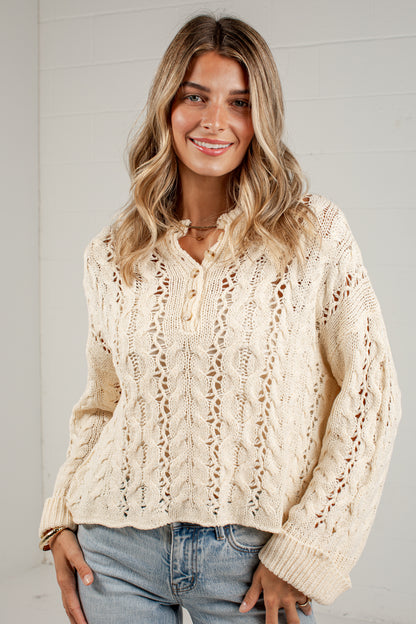 A woman wearing a cream cable-knit button-up sweater and light-wash blue jeans, standing against a white paneled wall.